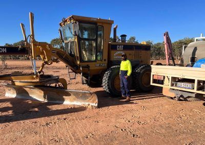 Man standing next to a yellow Caterpillar grader on a red dirt road. Sunny day.