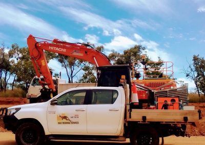 White work truck with orange excavator and equipment in an outdoor setting under a blue sky.
