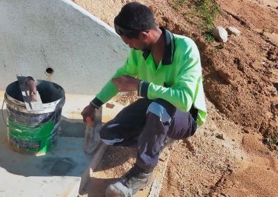 Person in green shirt using a trowel on concrete, next to a bucket. Outdoor setting, dirt slope in background.
