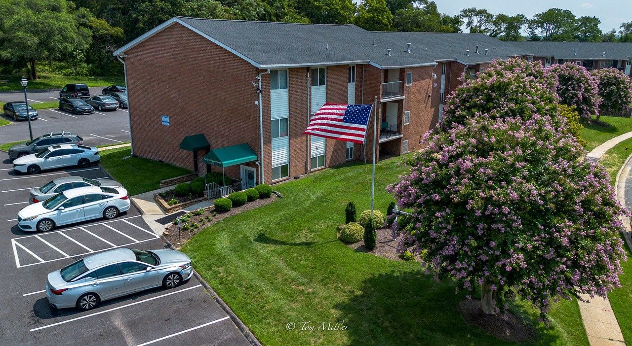 Aerial view of a brick apartment building with an American flag, parked cars, and a flowering tree by a curved road