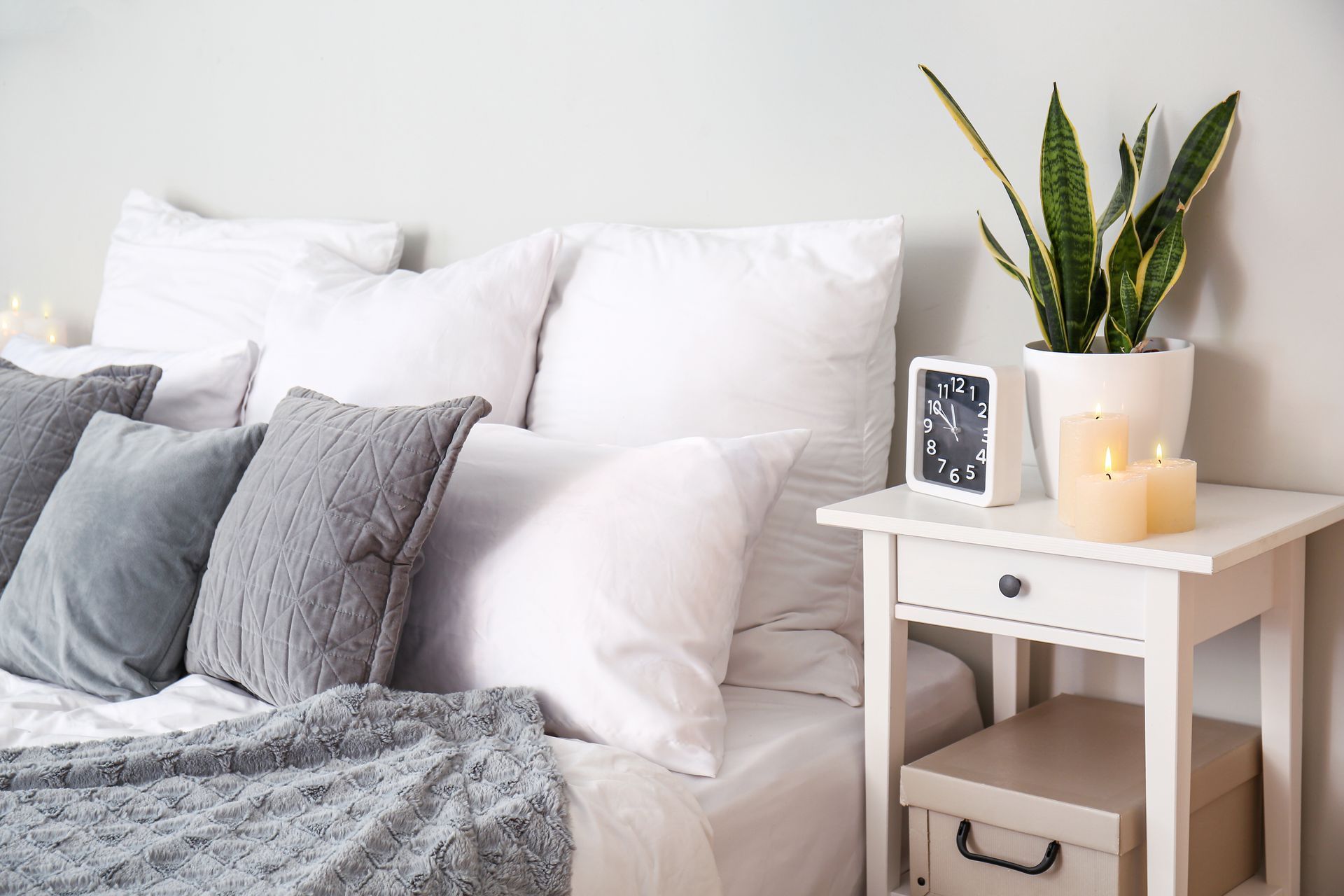 Cozy bedroom corner with white bed, gray pillows, and a bedside table with a plant and clock.