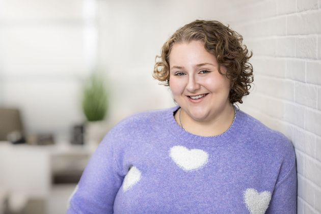 A woman in a purple sweater is leaning against a white wall and smiling.