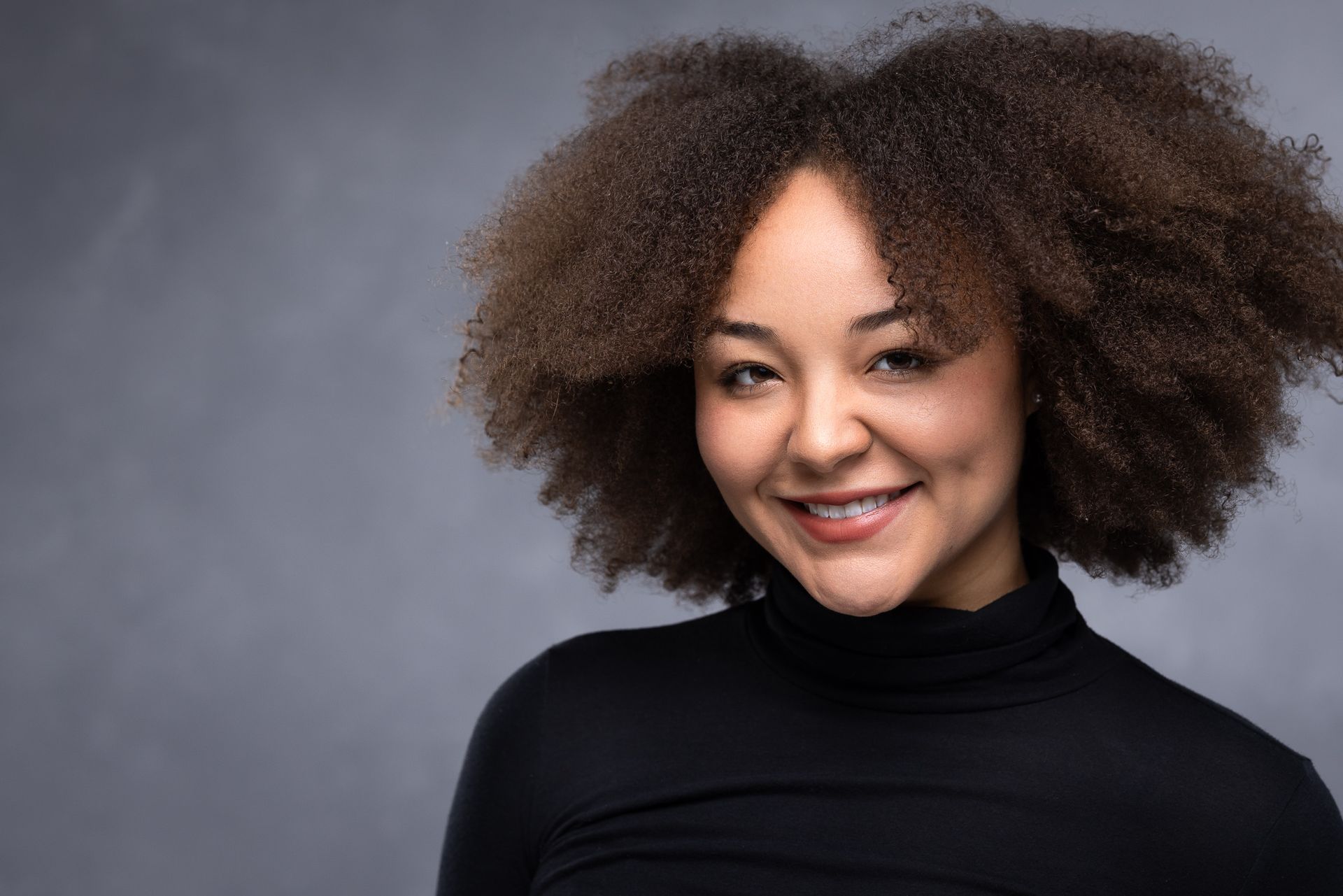 A woman with curly hair is wearing a black turtleneck and smiling.