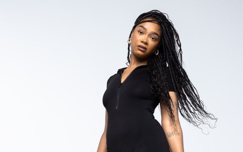 A woman in a black dress with braids is standing in front of a white background.