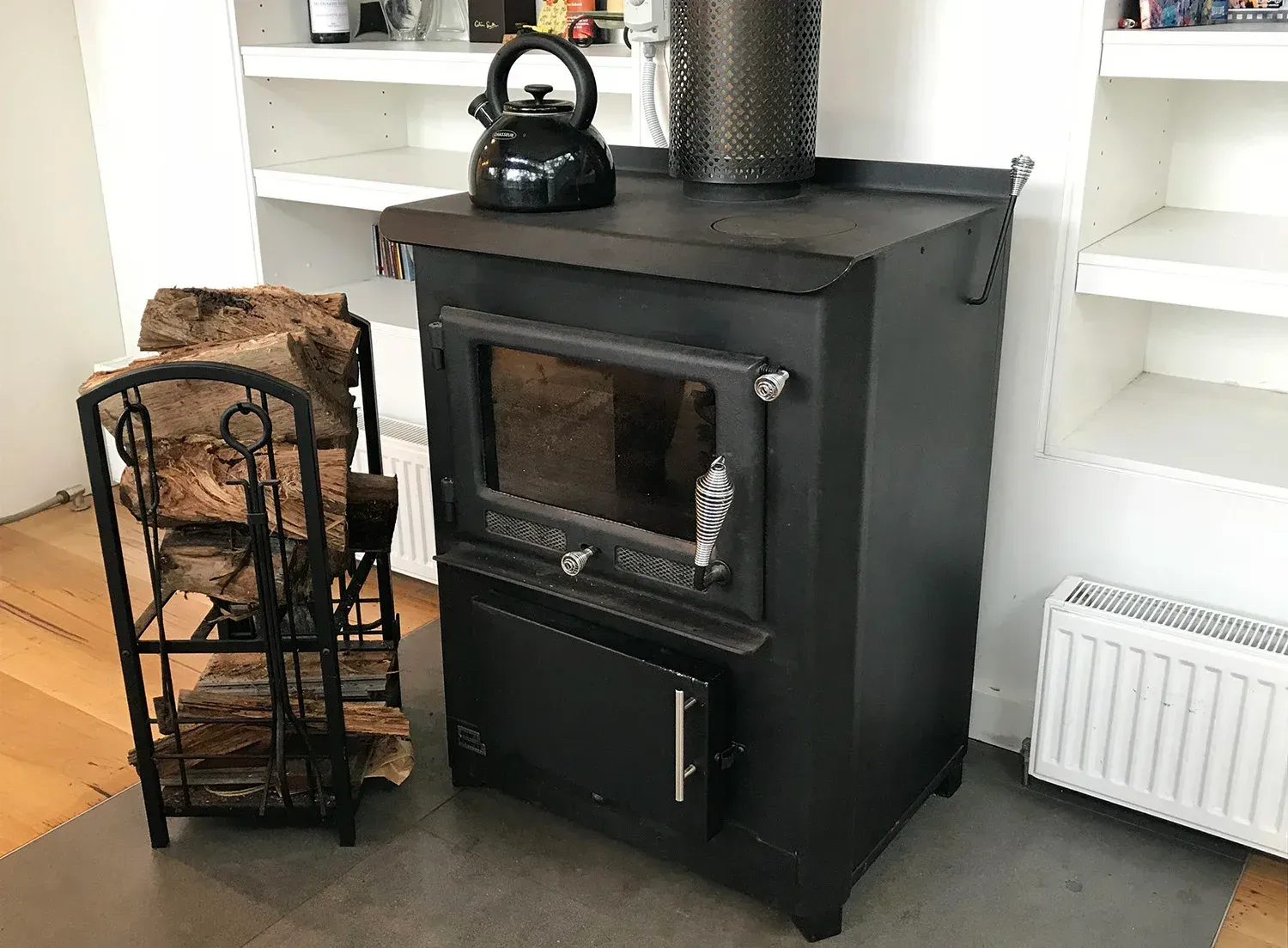 Black Wood Stove With Kettle, Beside a Basket of Logs in a White Built-in Shelving Alcove — Complete Heating In Huntingfield, TAS