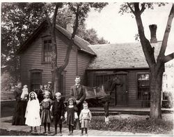 Children and a woman stand in front of a small wooden house with trees in the yard.