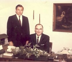 Two men in suits in an office, one seated at a desk and one standing behind him, with flowers in front.