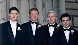 Four men in tuxedos standing together at a formal event, with white boutonnieres.
