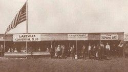 People standing outside Lakeville Commercial Club storefronts with signs, including American and Supreme Gas