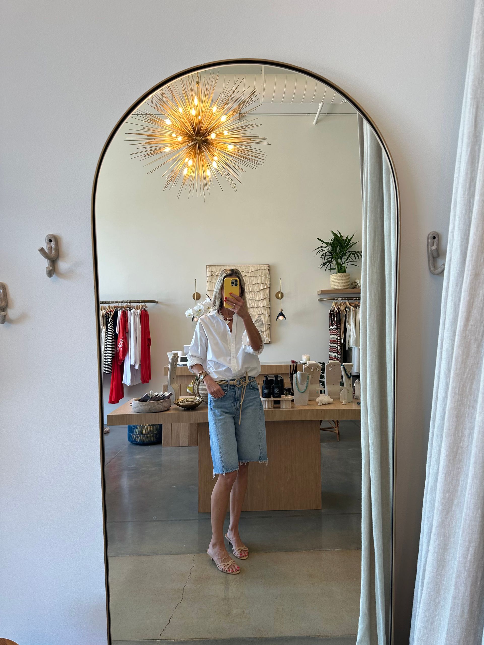 Person taking a mirror selfie in a bright room with a starburst chandelier, desk, and plant.