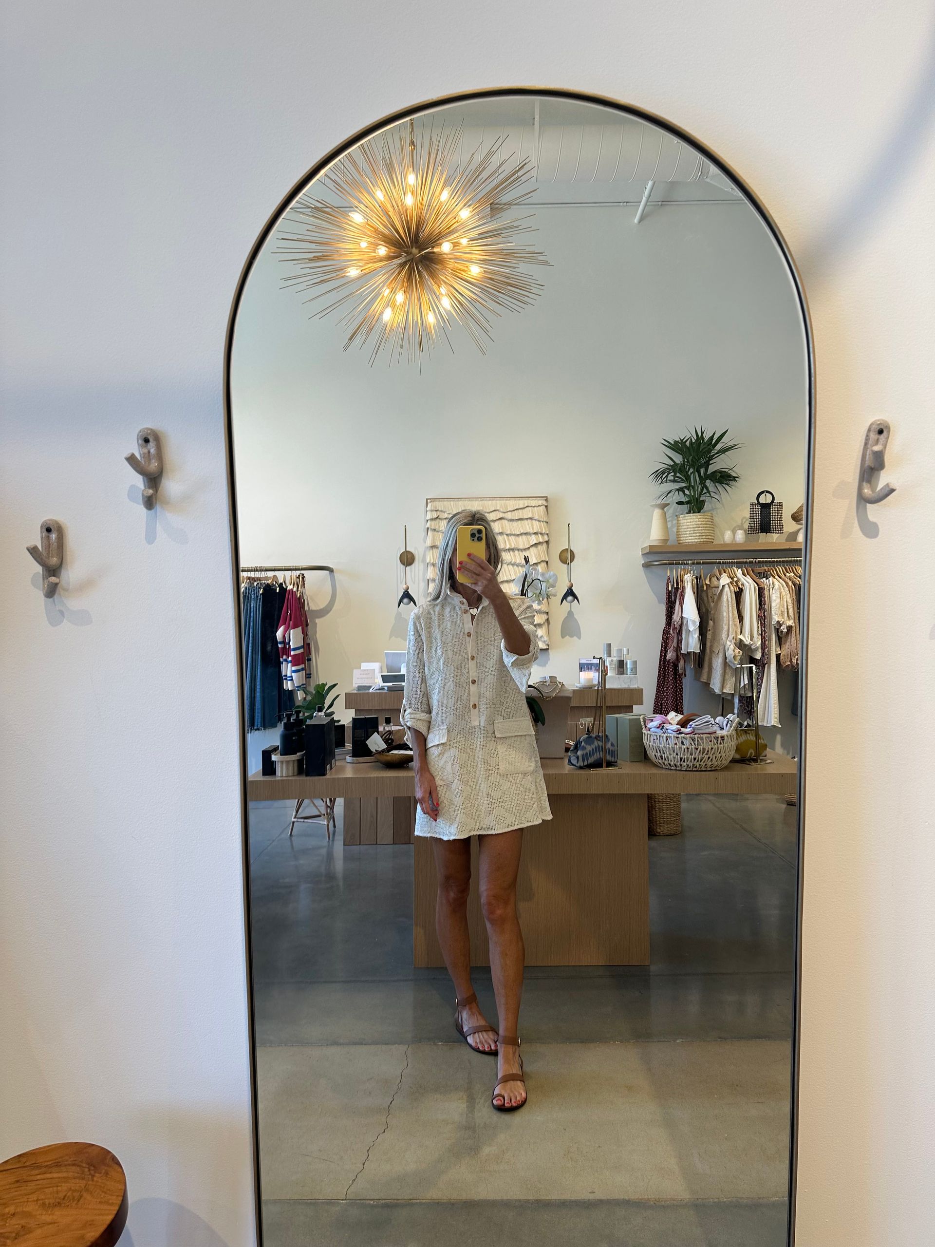 Woman taking a mirror selfie in a boutique, wearing a white dress and heels under a starburst light.