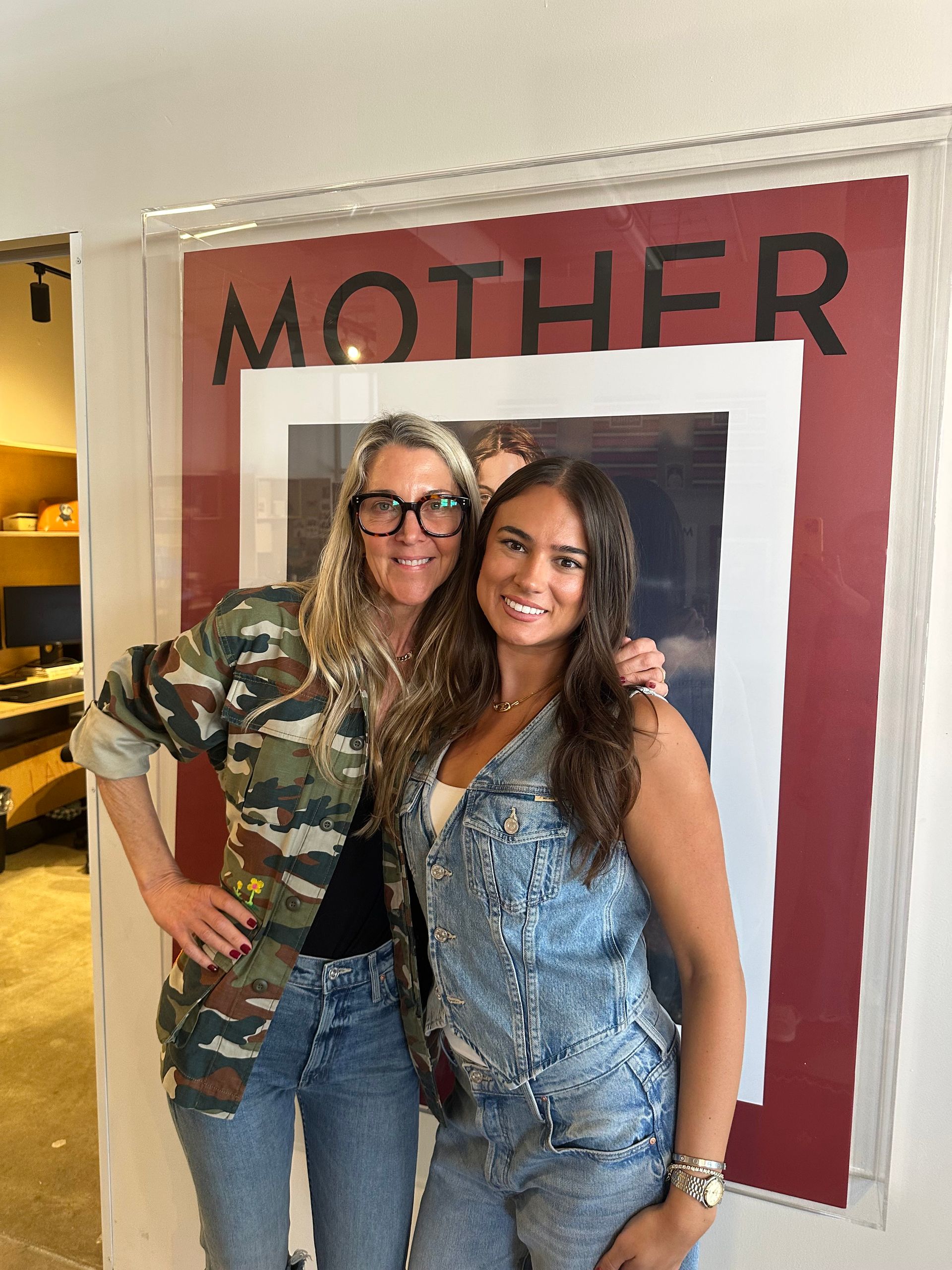 Two women posing in front of a red “MOTHER” backdrop, smiling in a hallway