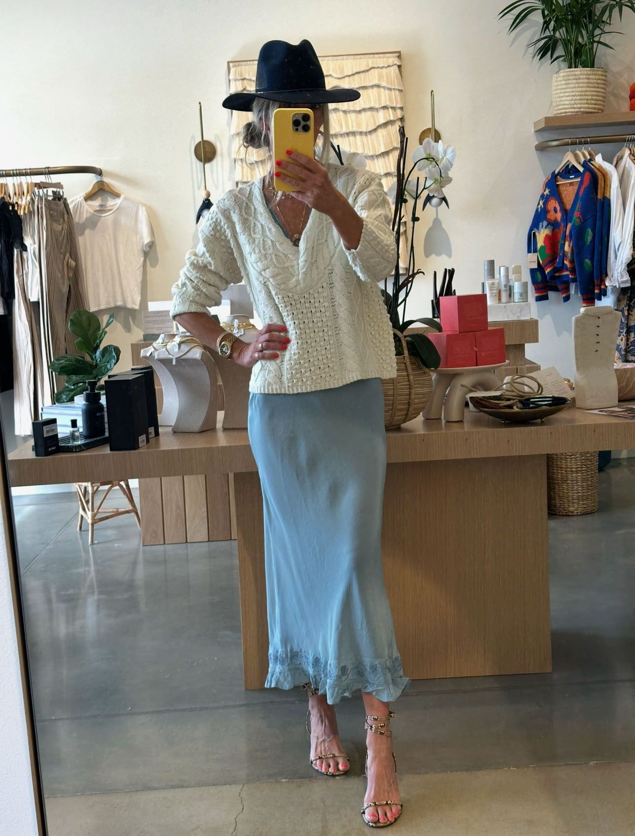 Person posing in a clothing store mirror, wearing a hat, white top, and light blue skirt.