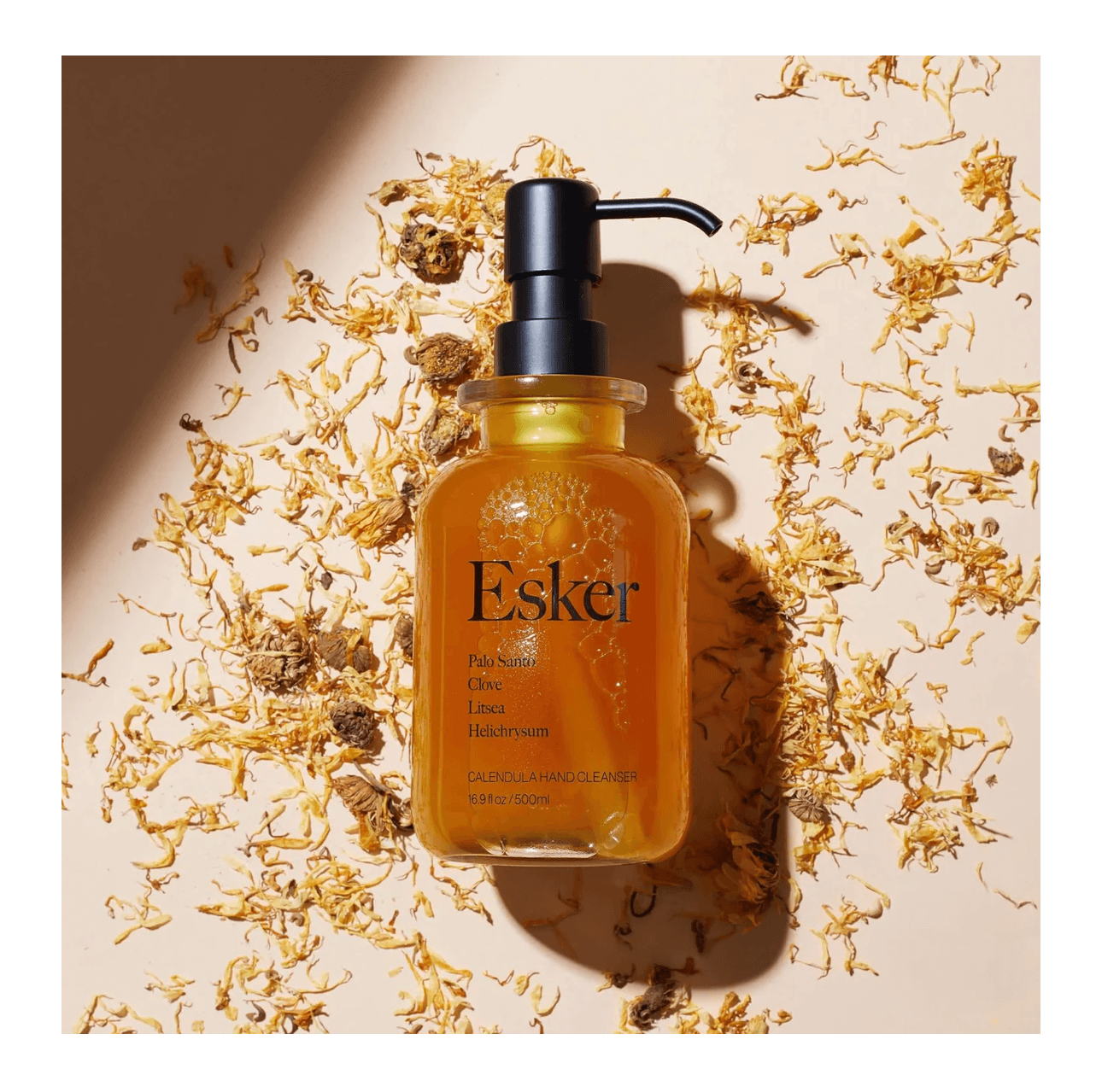 Amber Esker pump bottle on a beige background with scattered dried flowers and shadow.
