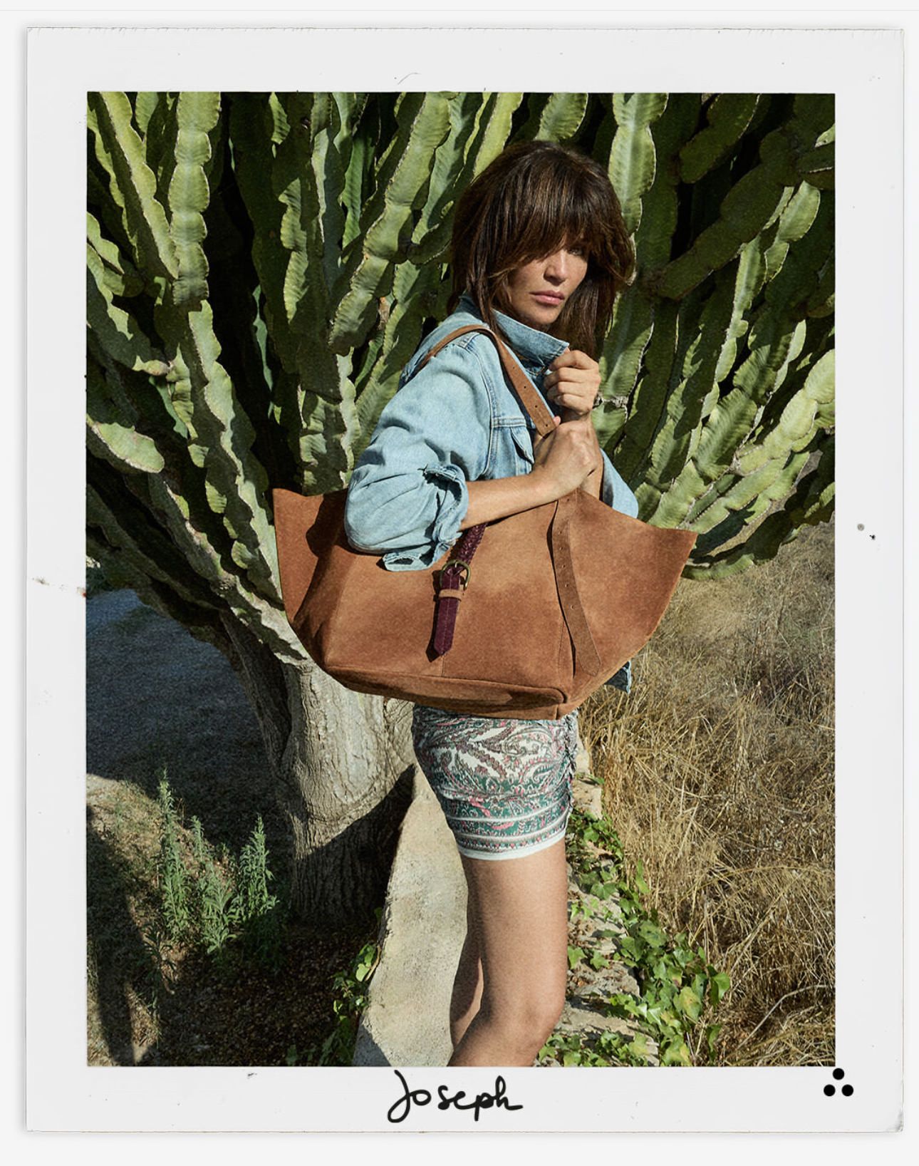 Woman posing outdoors in a denim jacket and patterned shorts, holding a large tan tote by cactus plants.
