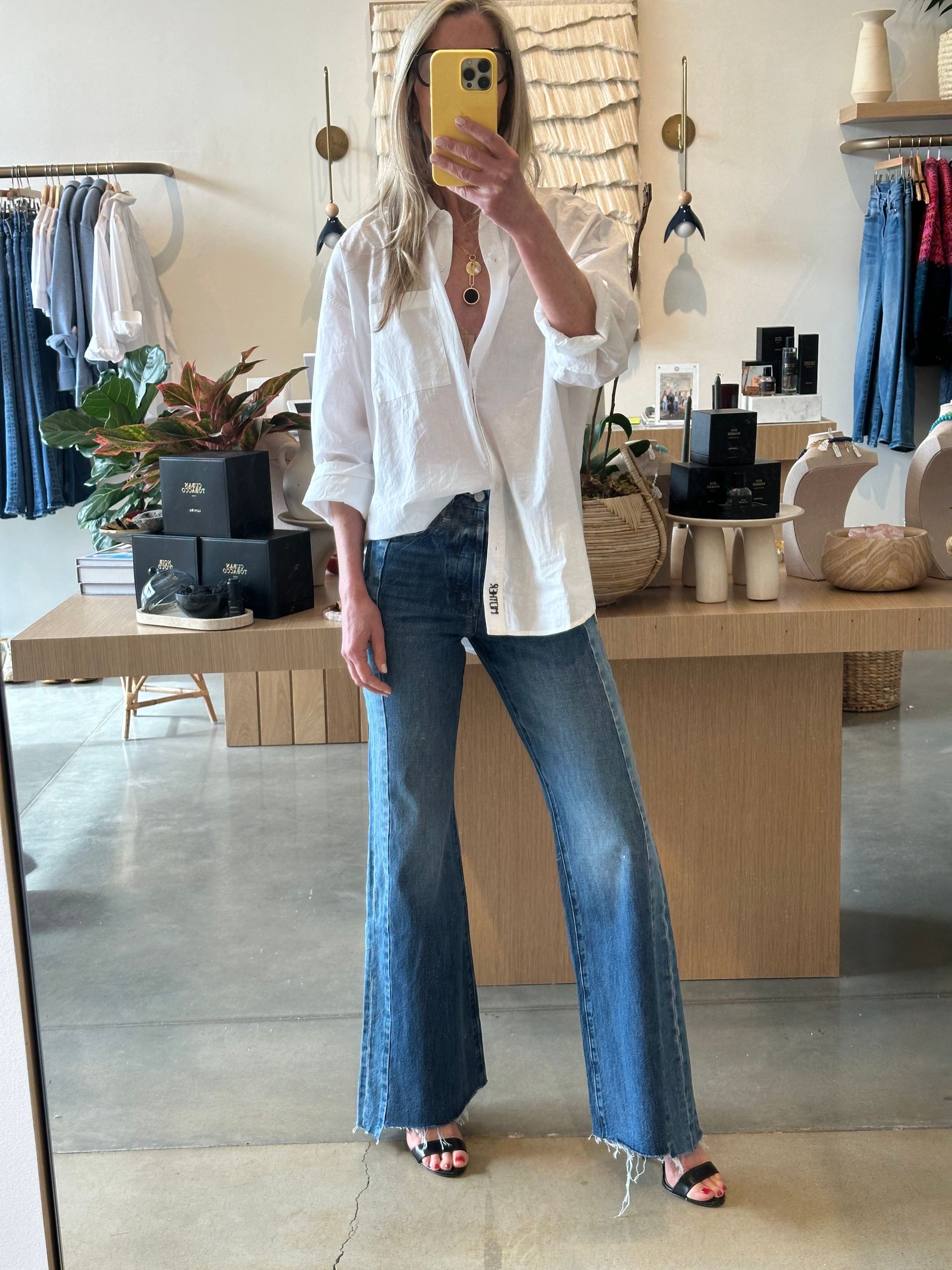 Person taking a mirror selfie in a clothing boutique, wearing a white shirt, blue jeans, and heels.