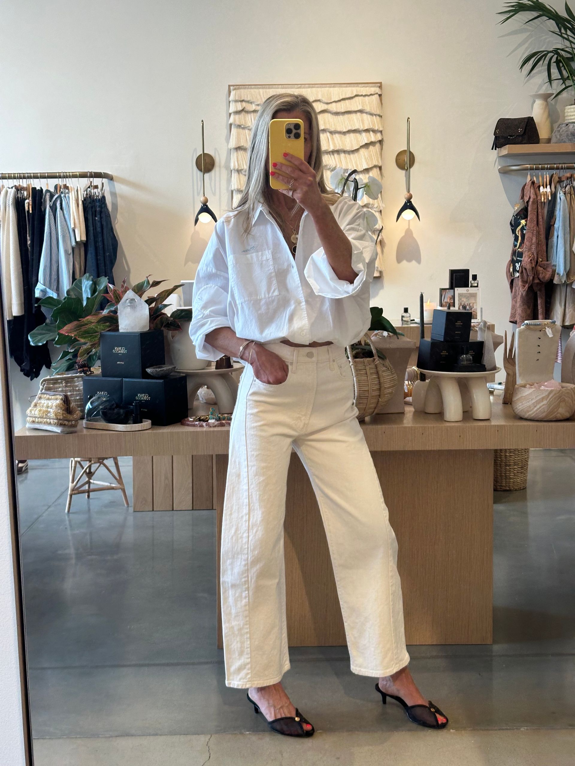 Person taking a mirror selfie in a boutique, wearing a white blouse, cream pants, and black heels.