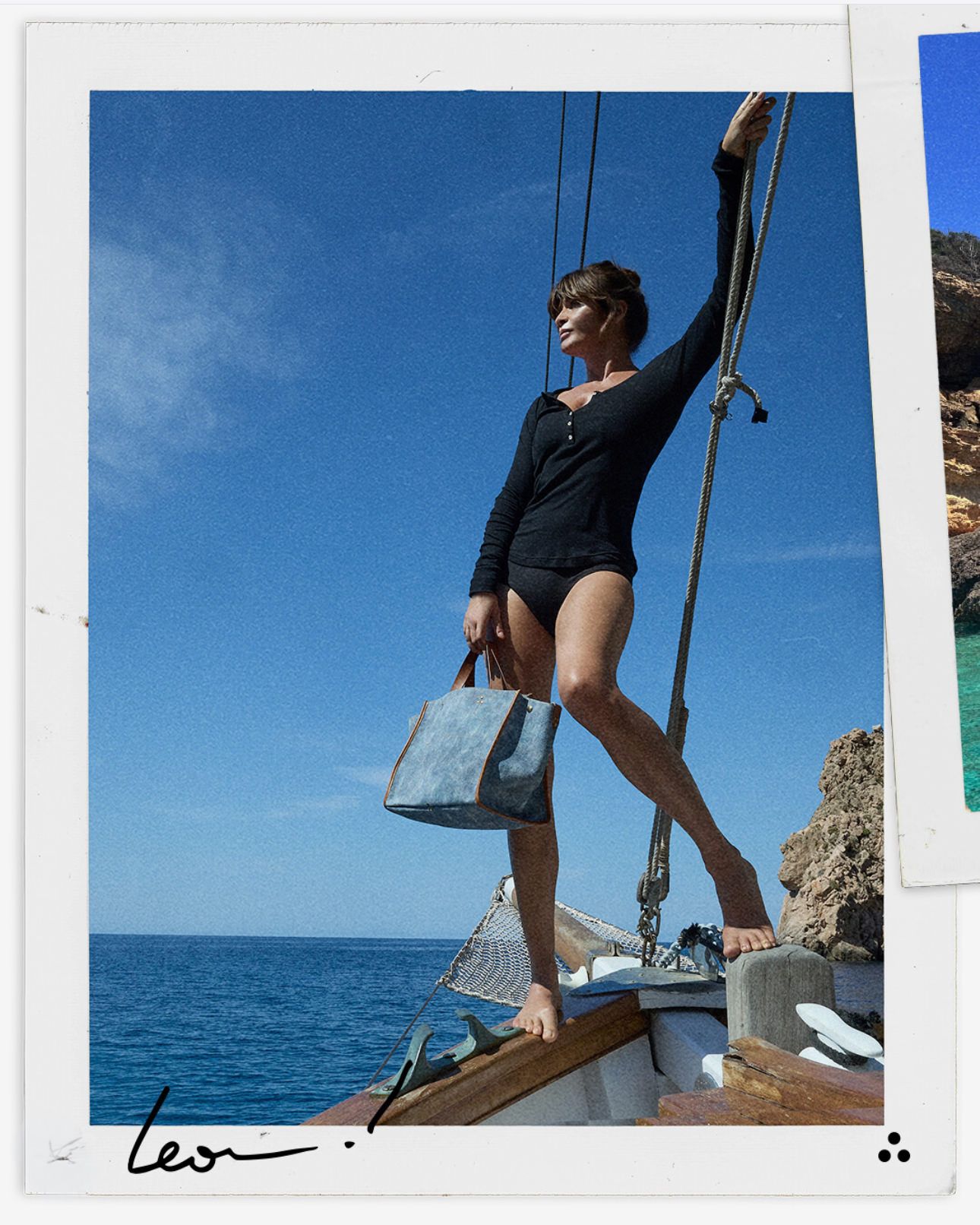 Woman standing on a sailboat in a black swimsuit under a clear blue sky, holding a blue bag.