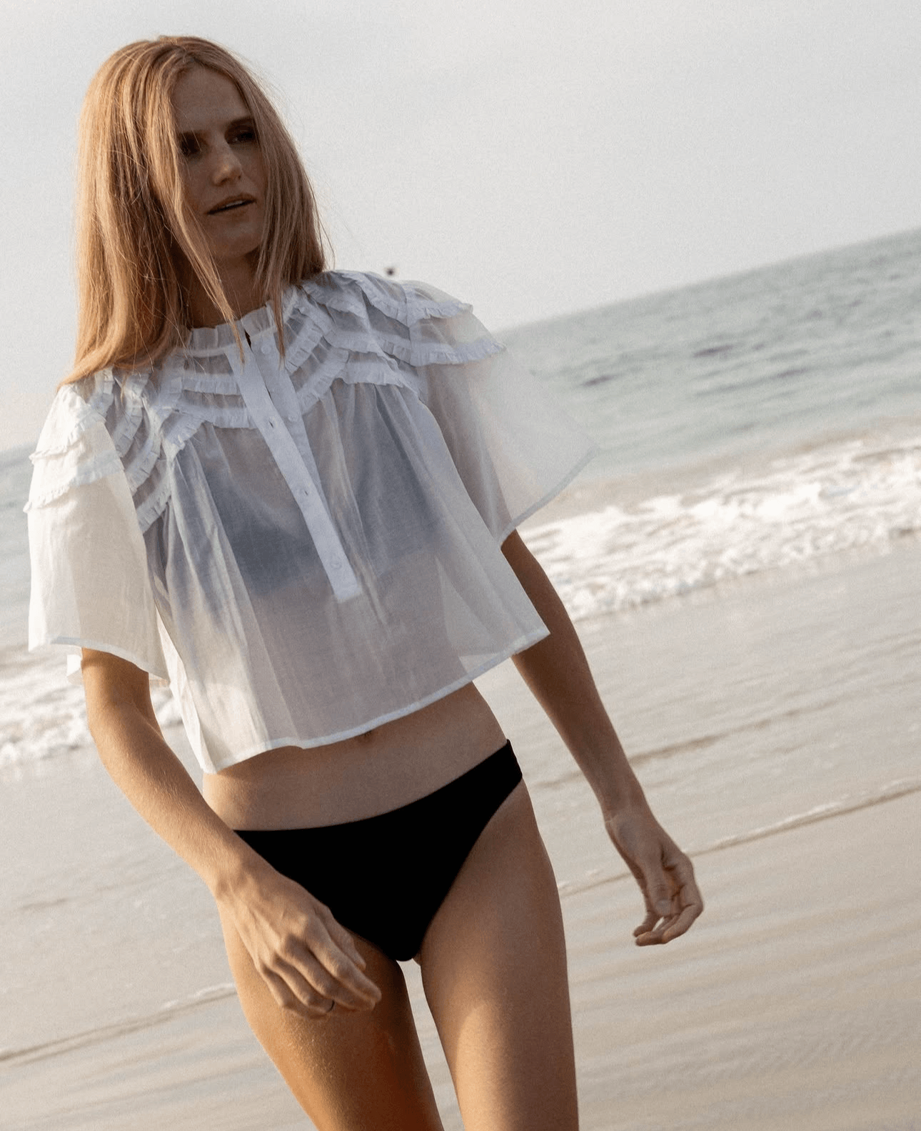Person walking on a beach in a white sheer top and black bottoms, with ocean waves in the background