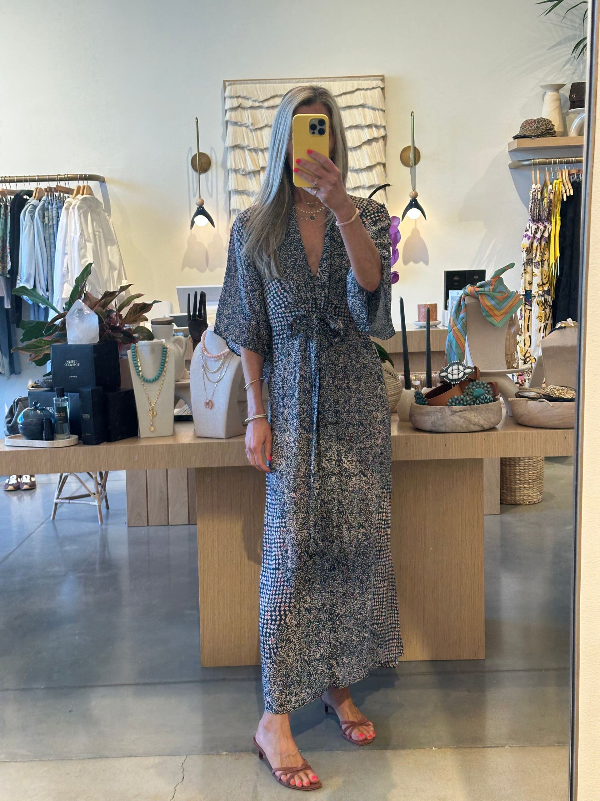 Person in a blue patterned dress taking a mirror selfie in a clothing boutique.