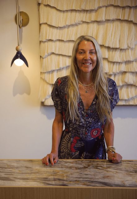 Woman smiling behind a marble counter in a softly lit room with textured wall art.