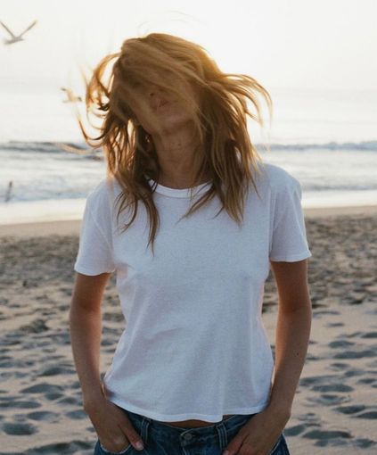 Person with messy hair standing on a beach in a white T-shirt and jeans, ocean in the background