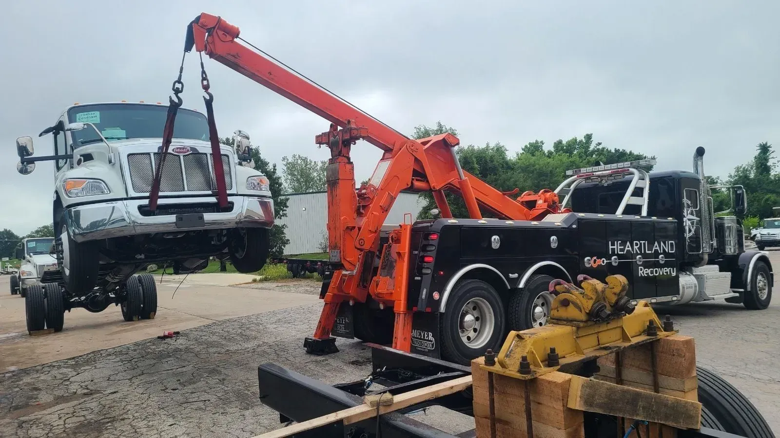 An orange heavy-duty wrecker truck lifts the front end of a white semi-truck via tow straps in an outdoor gravel lot.