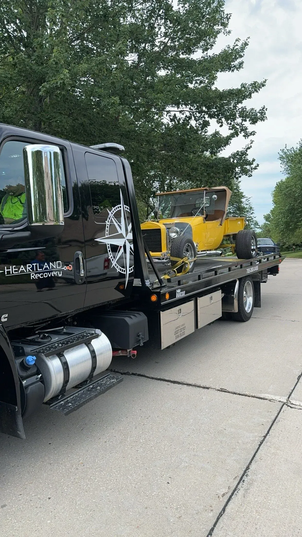 A black tow truck transporting a yellow vintage roadster on a paved road lined with trees.