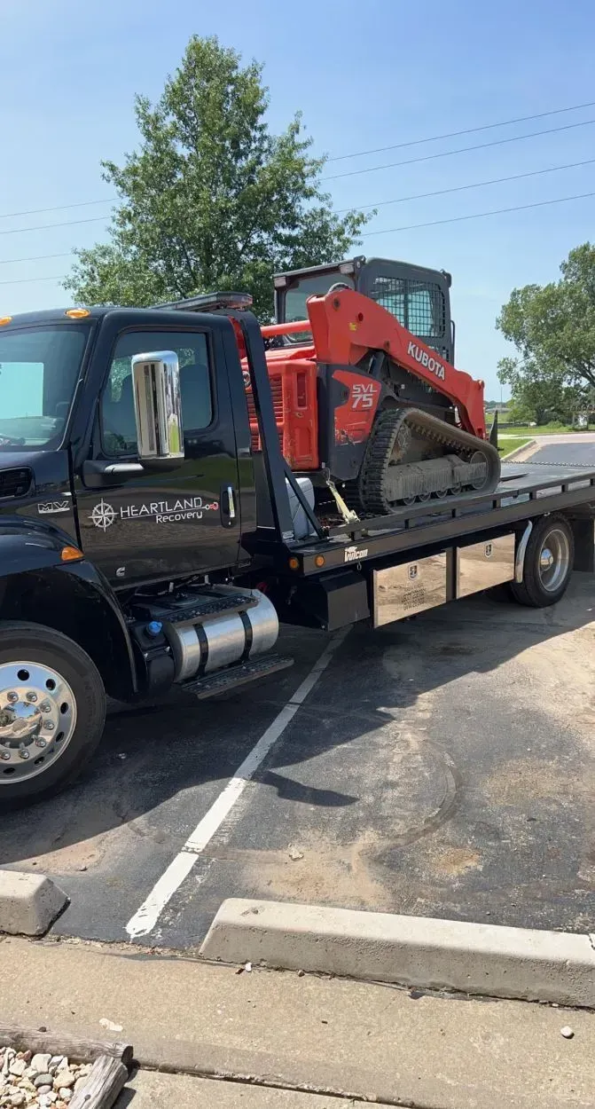 A black flatbed tow truck carrying an orange skid steer loader parked in a paved lot.