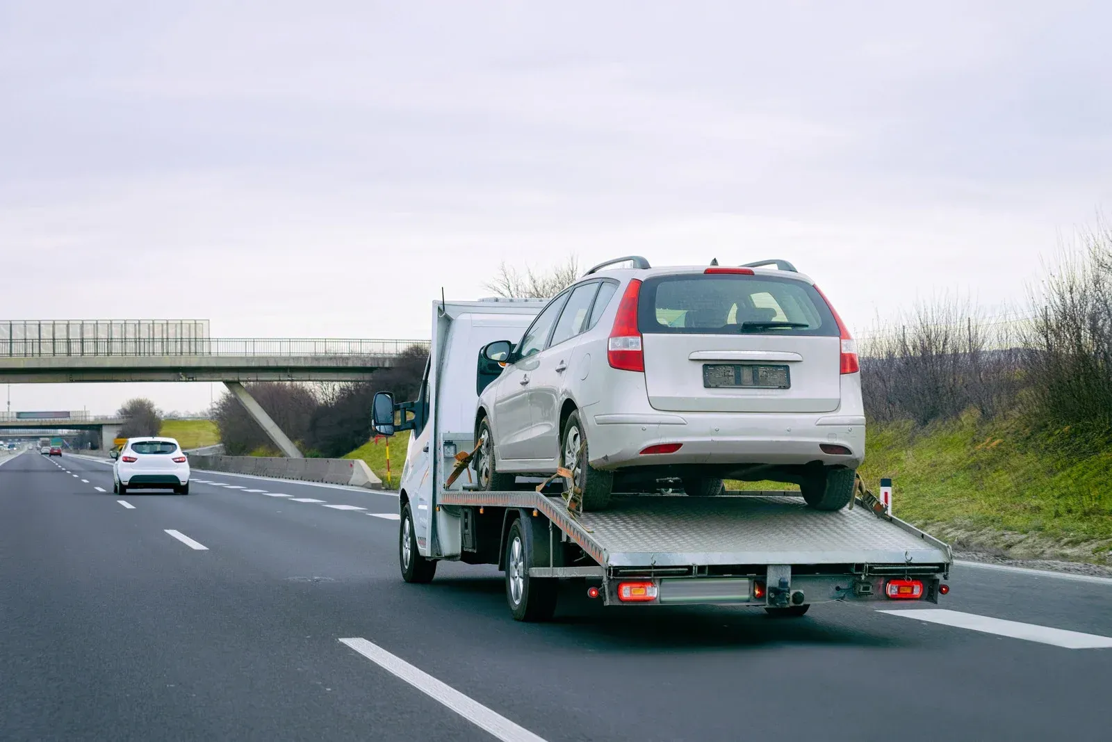 A white tow truck transports a silver station wagon on a multi-lane highway under an overcast sky.