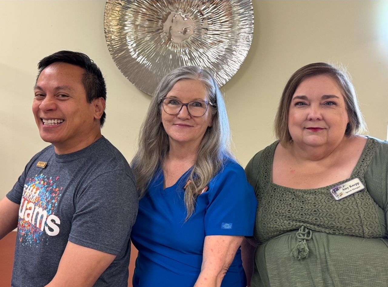 Three people smiling, indoors. Man in grey shirt, woman in blue scrubs, woman in green dress.