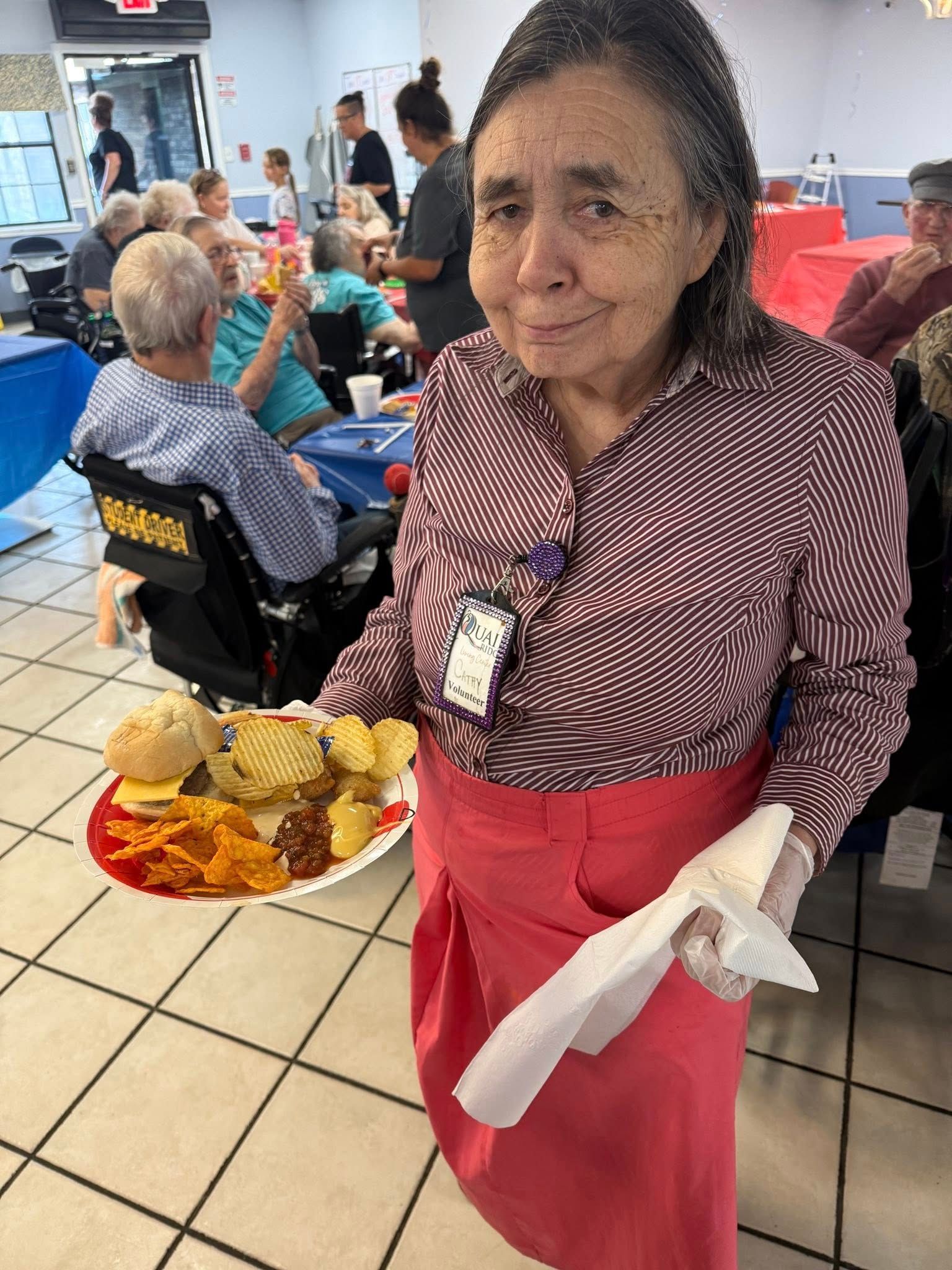 Woman in red apron holds a plate of food, smiling at the camera. Other people sit at tables in the background.