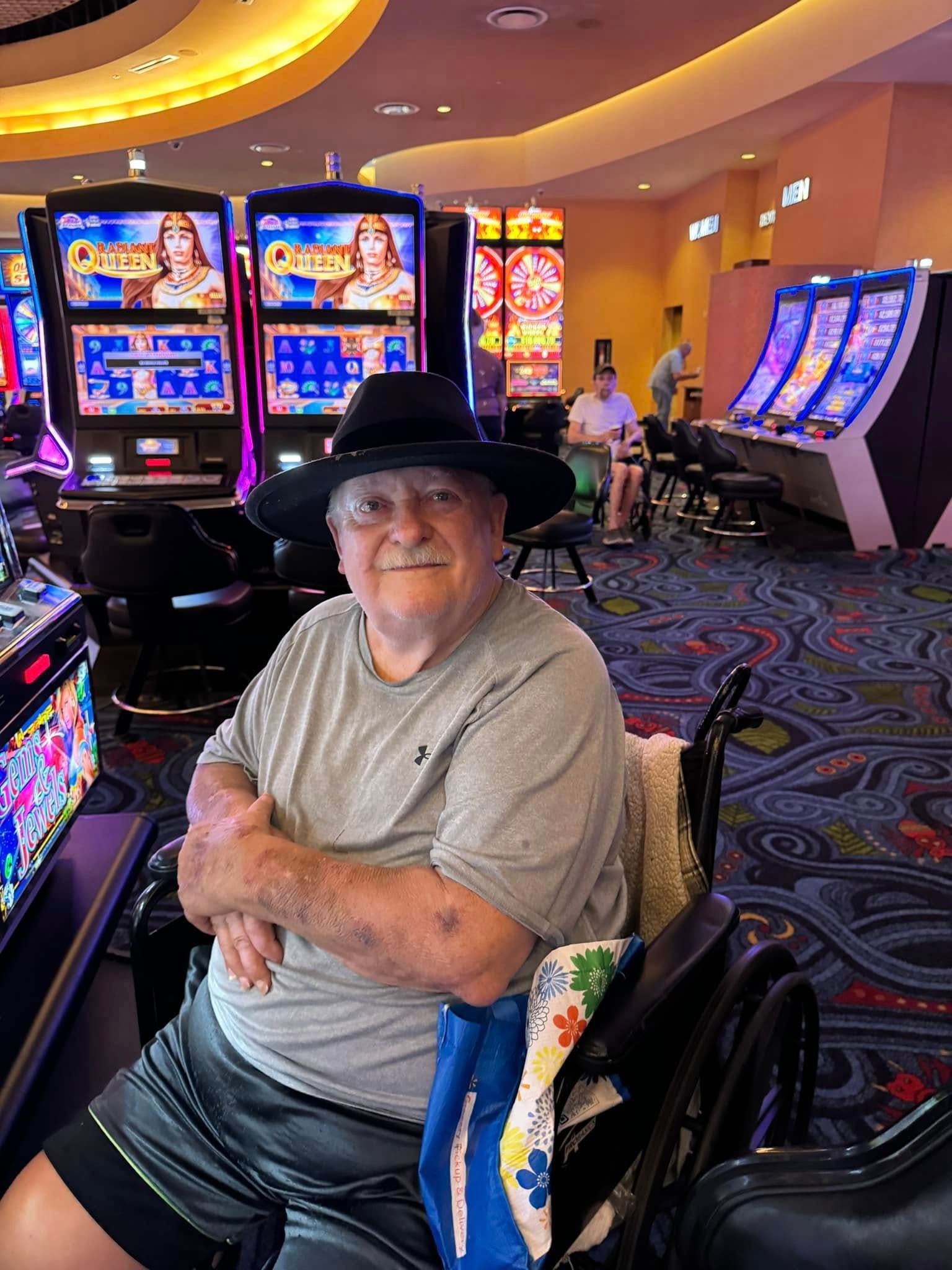An older man in a wheelchair wearing a hat, sits in a casino, smiling.