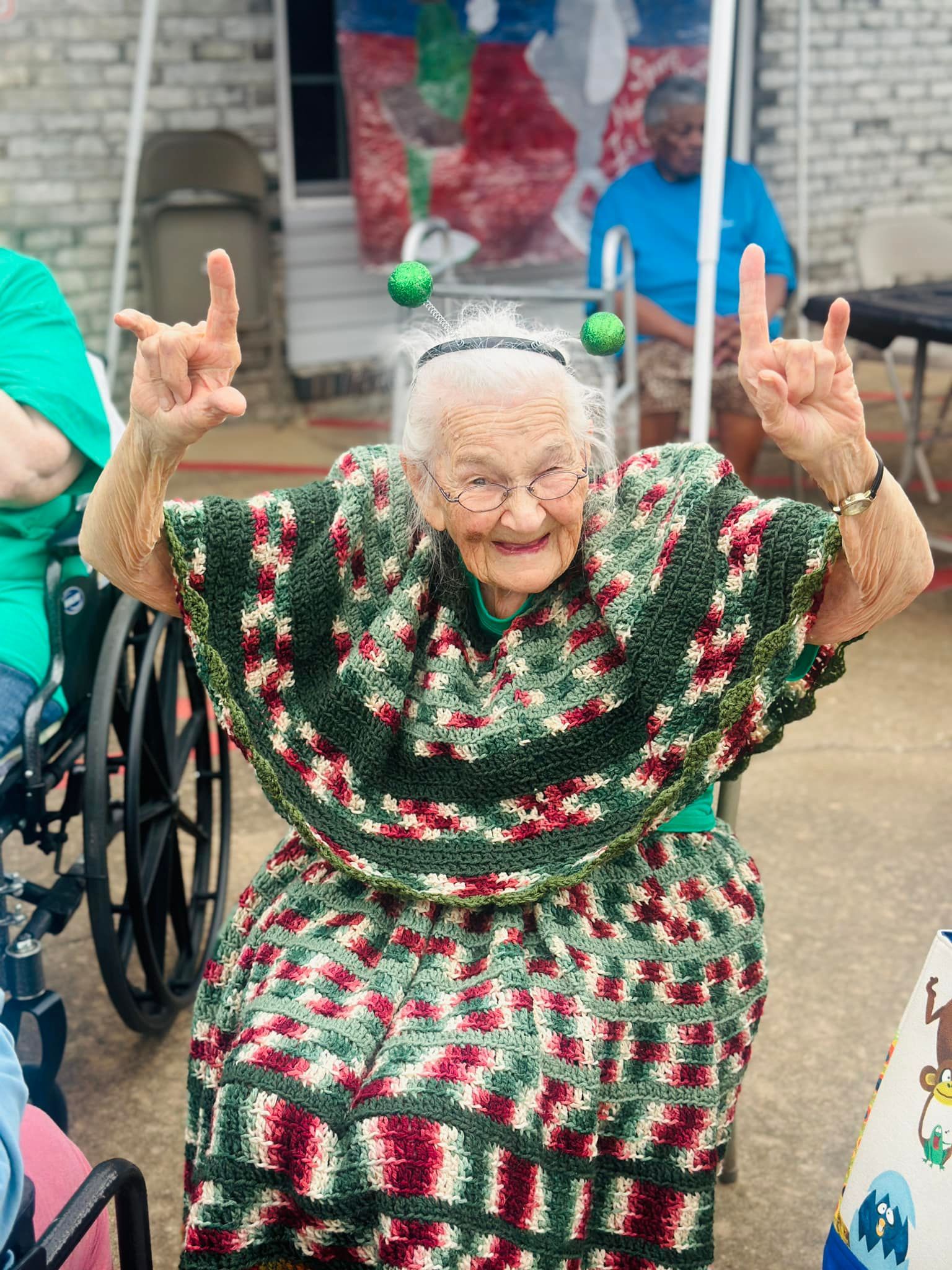 Elderly woman in wheelchair making rock on hand gesture, wearing a colorful knitted outfit and alien headpiece.