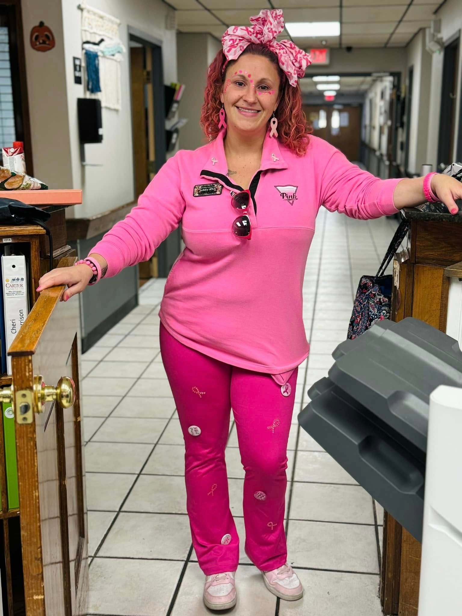 Woman in bright pink outfit stands in hallway, smiling. She wears a bow, jewelry, and carries a bag.