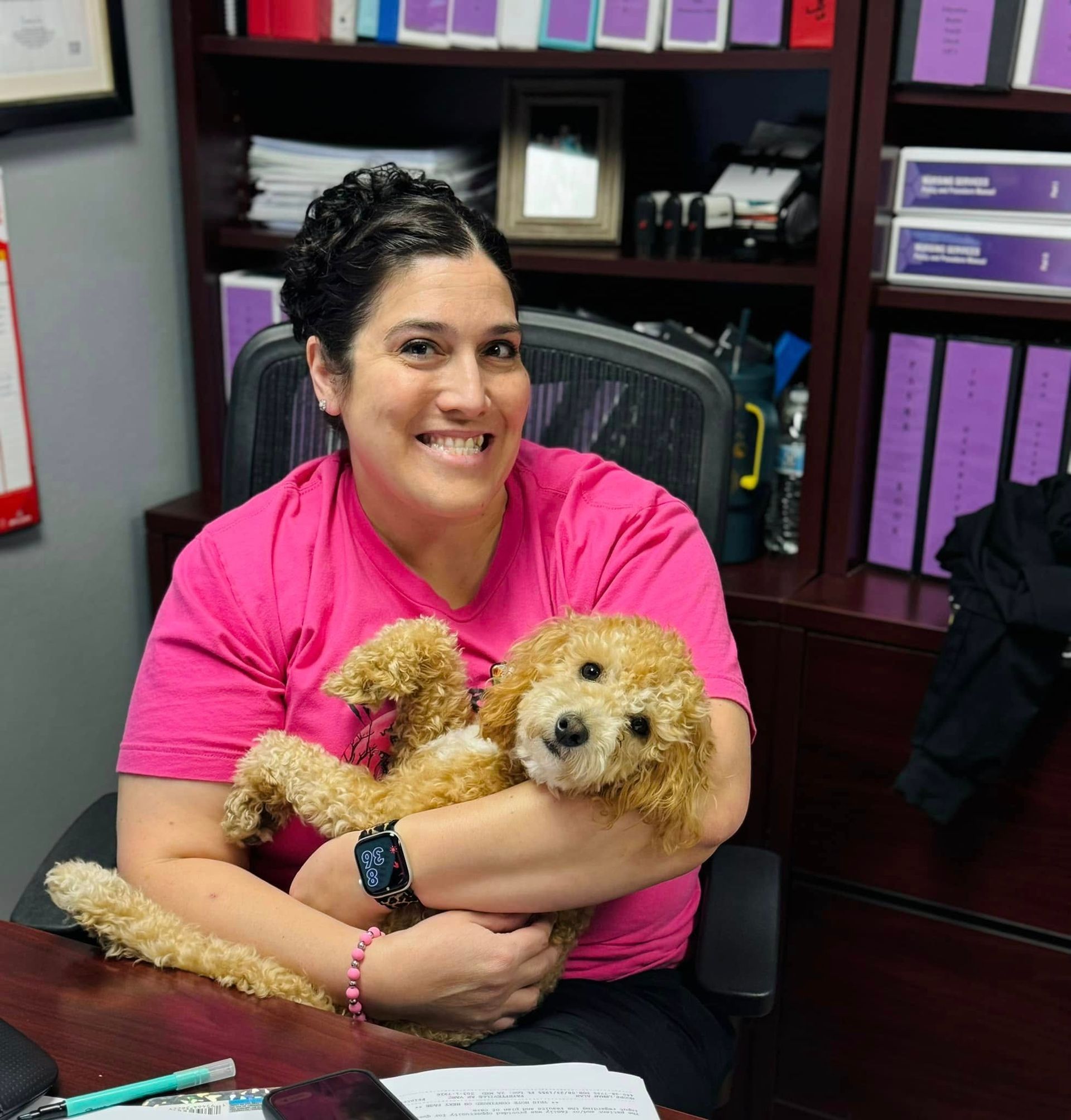 Woman in pink shirt smiles, holding a golden doodle puppy in an office setting with shelves and files.