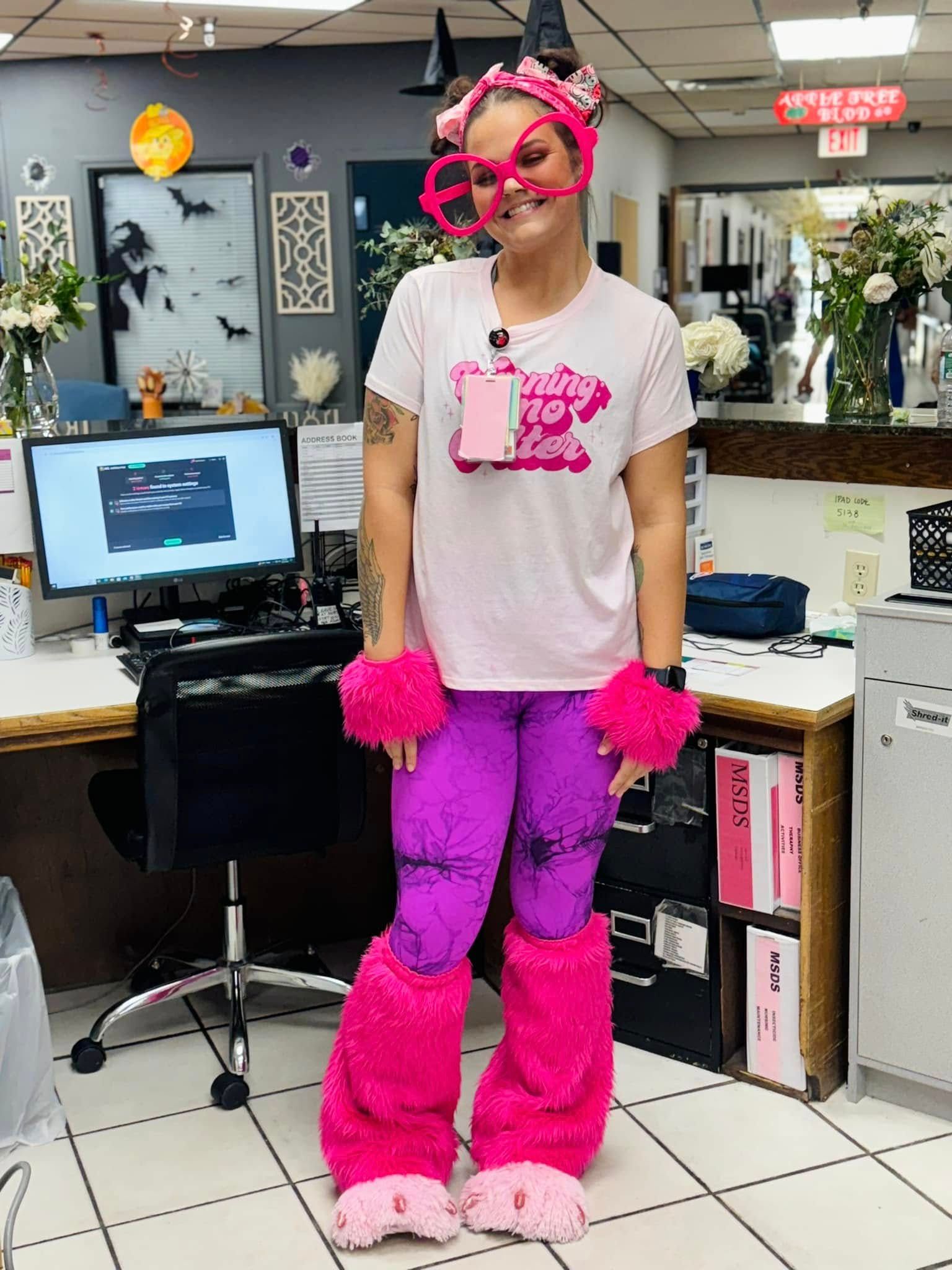 Woman in pink monster costume at a desk, with purple leggings and fuzzy cuffs.