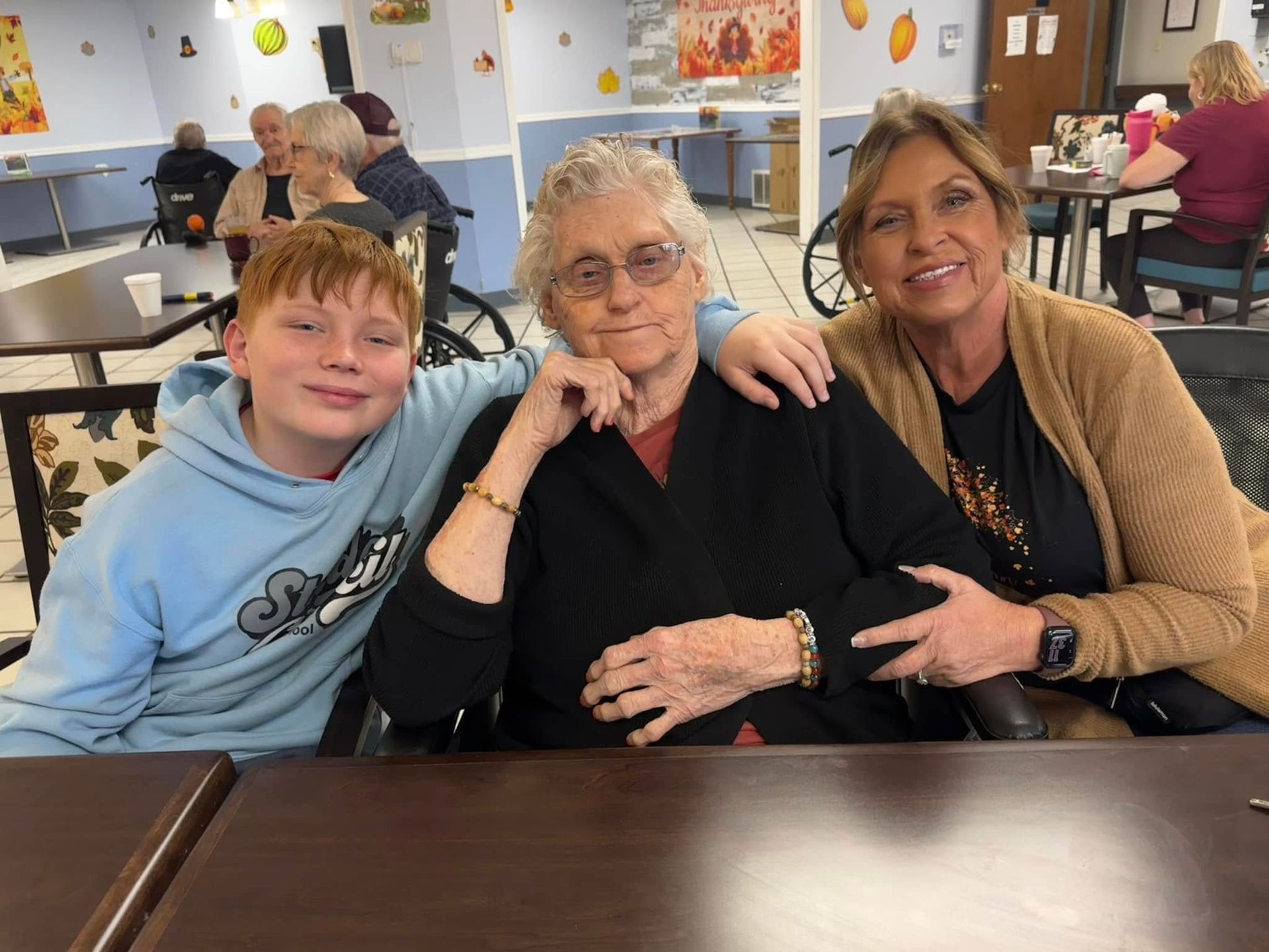 A boy, elderly woman, and woman smile together at a table. They are in a dining hall decorated for fall.