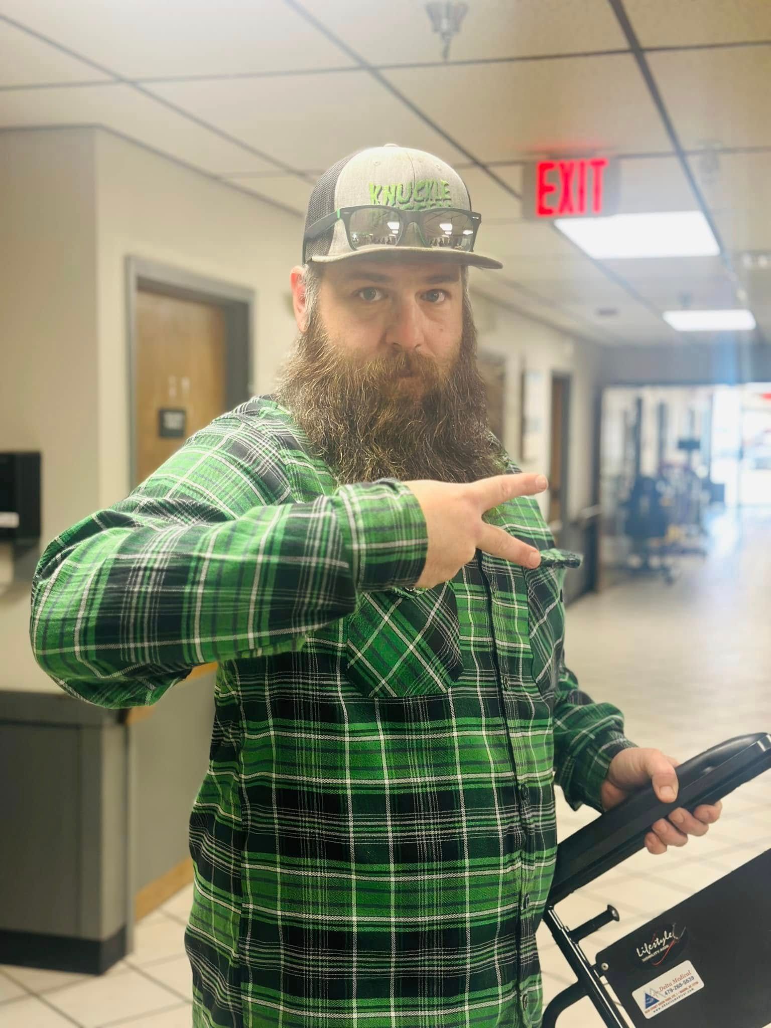 Man with beard in green plaid shirt, holding chair in hallway, giving peace sign.