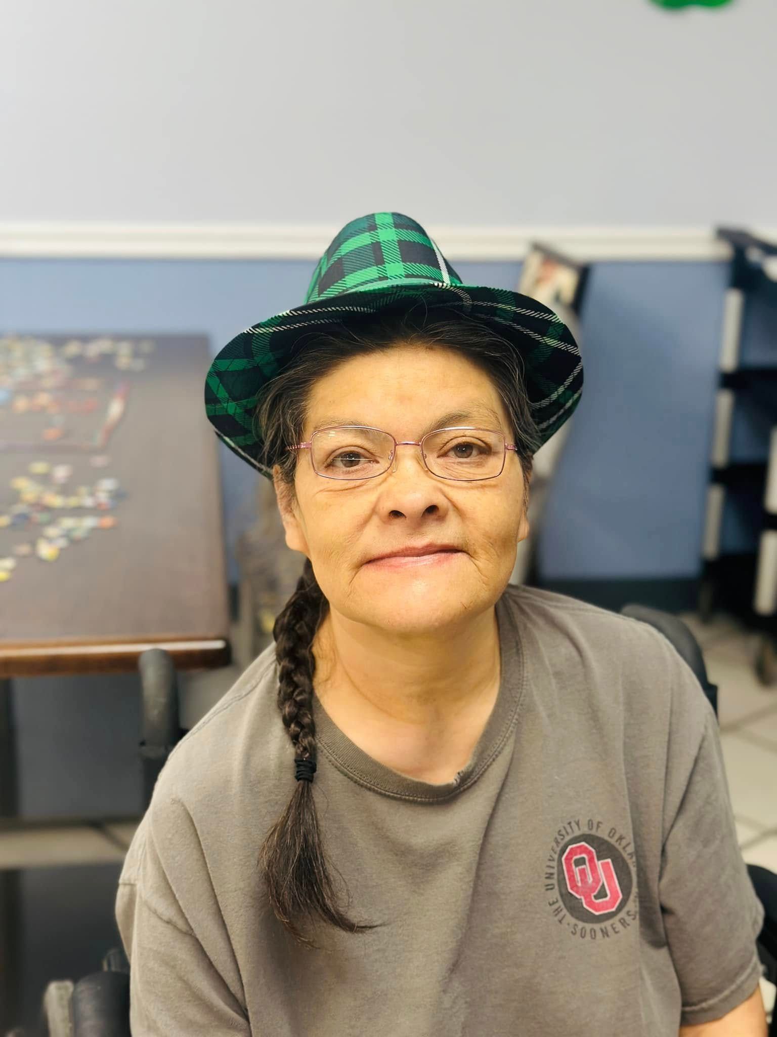 Woman with glasses and a green plaid hat, wearing a grey t-shirt, indoors near a puzzle.