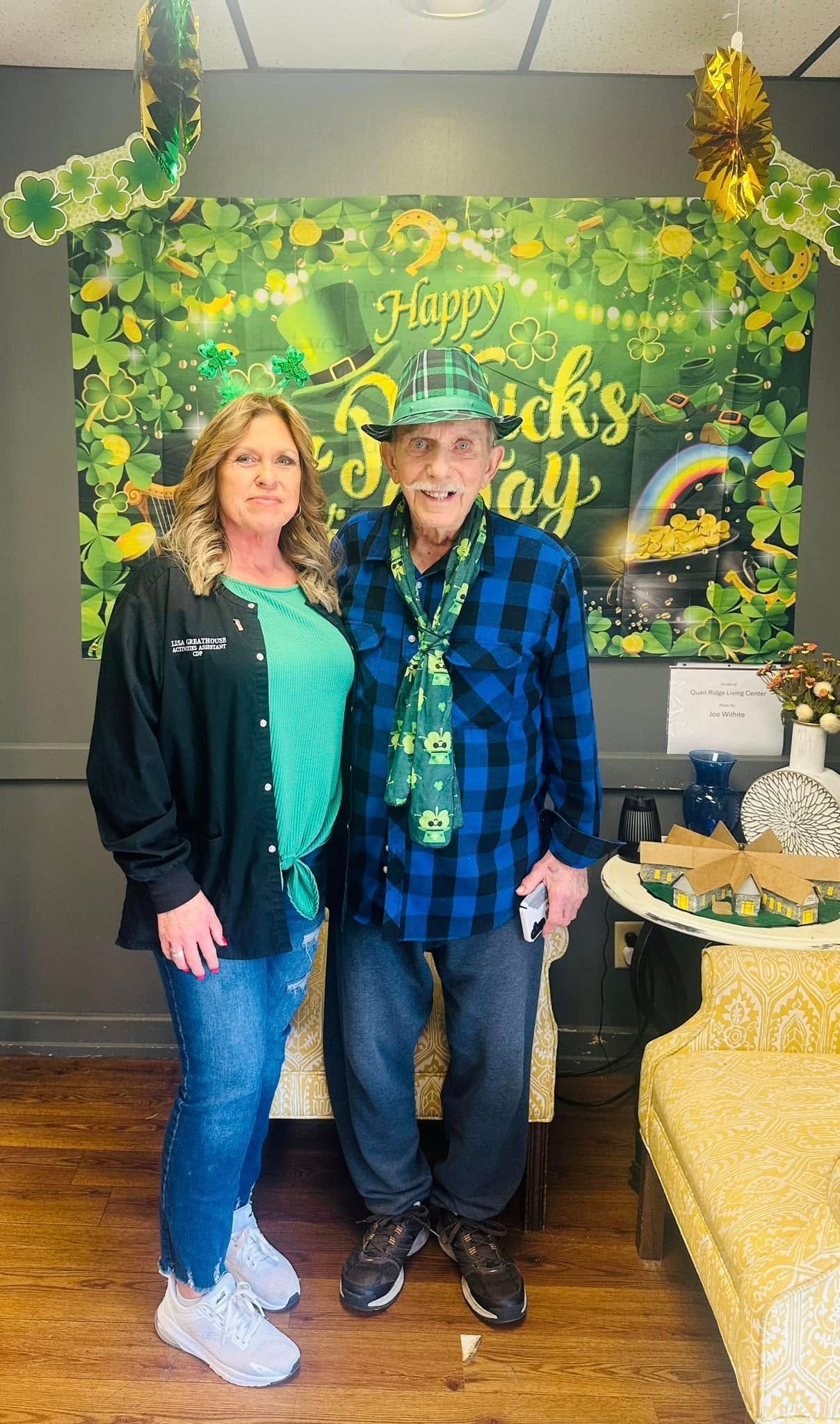 Woman and elderly man in St. Patrick's Day attire posing in front of a festive backdrop.