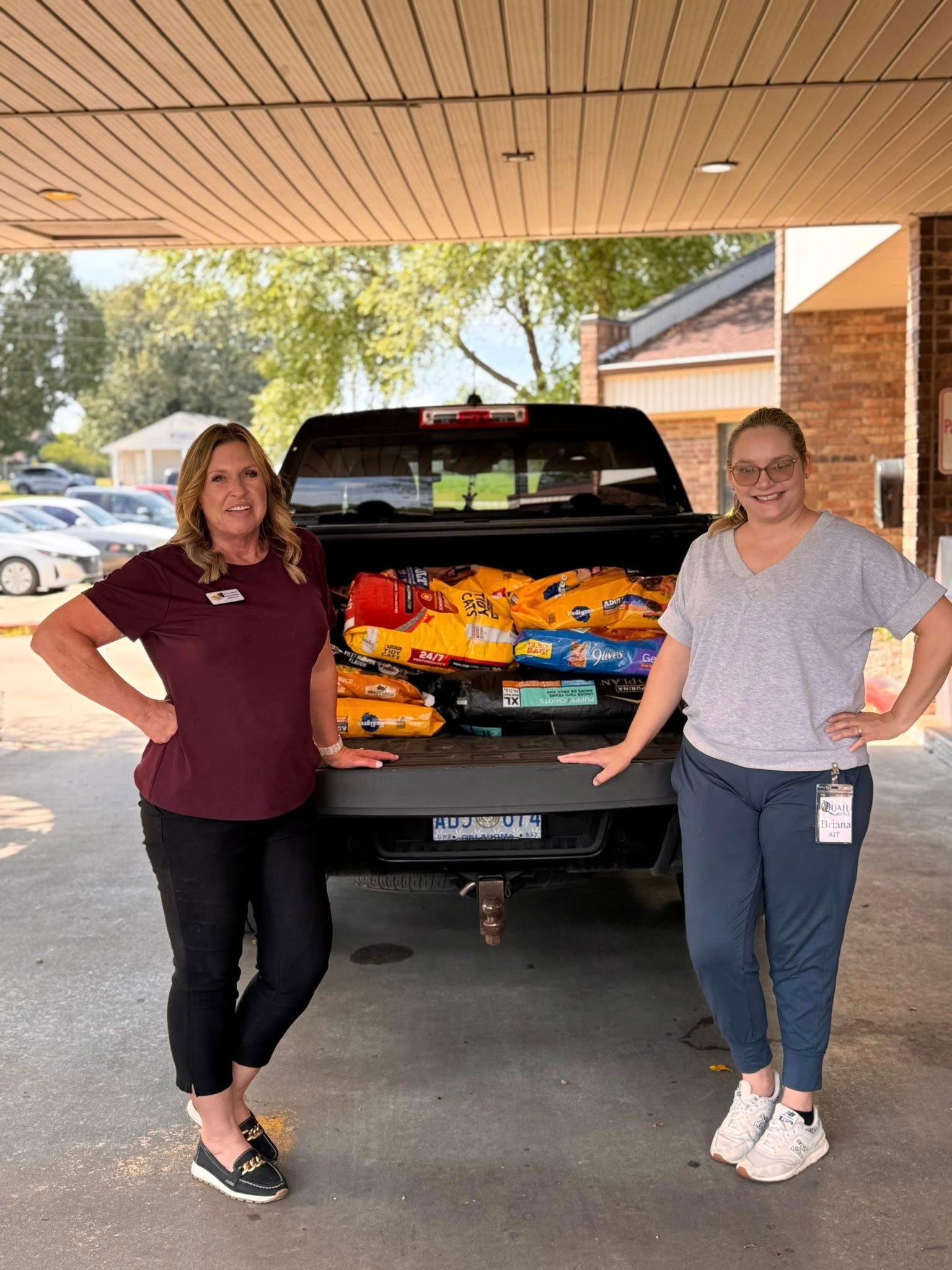 Two women pose with hands on hips beside a truck filled with pet food.