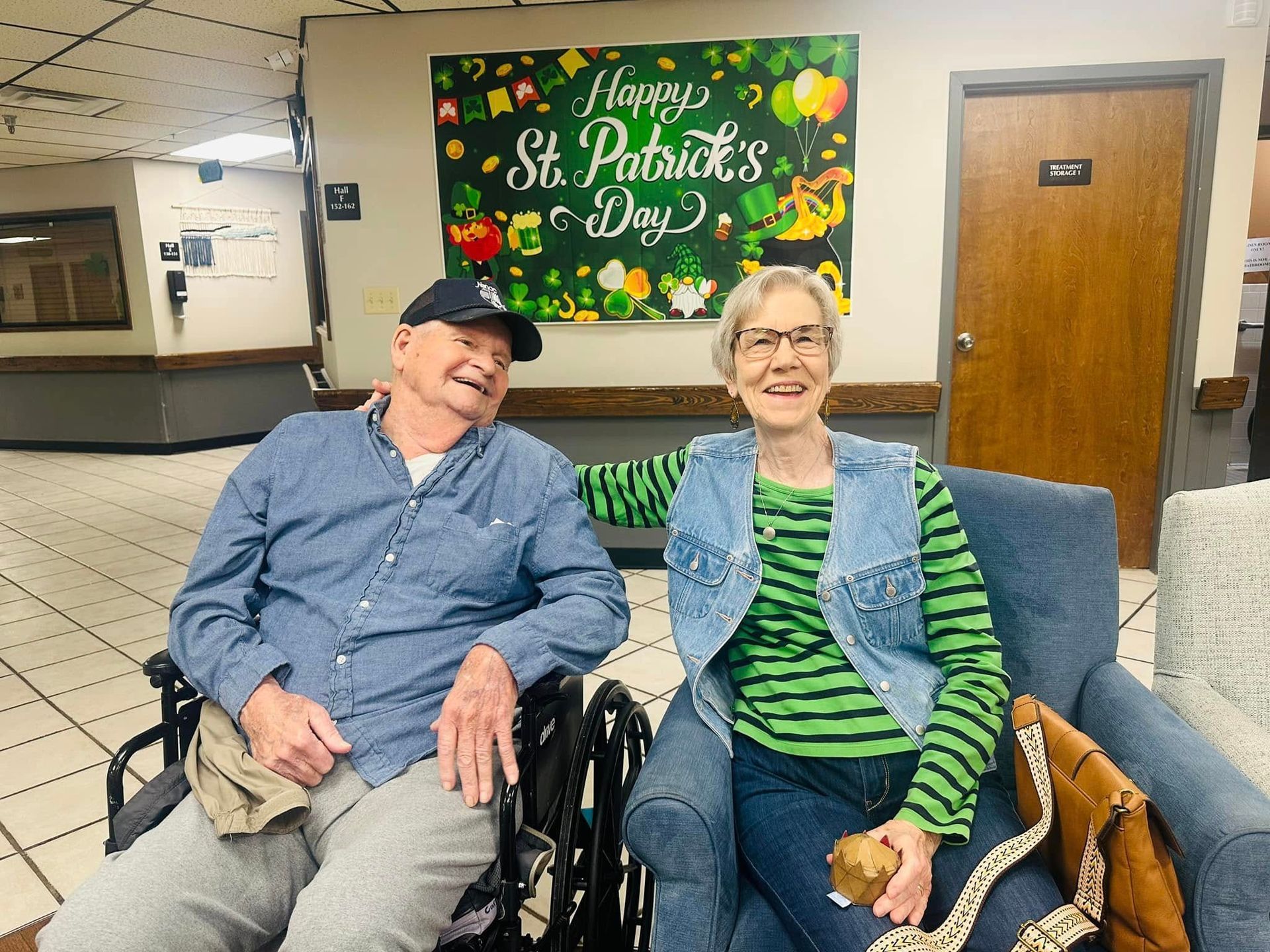 Two smiling seniors, one in a wheelchair, celebrate St. Patrick's Day. A green banner decorates the wall.