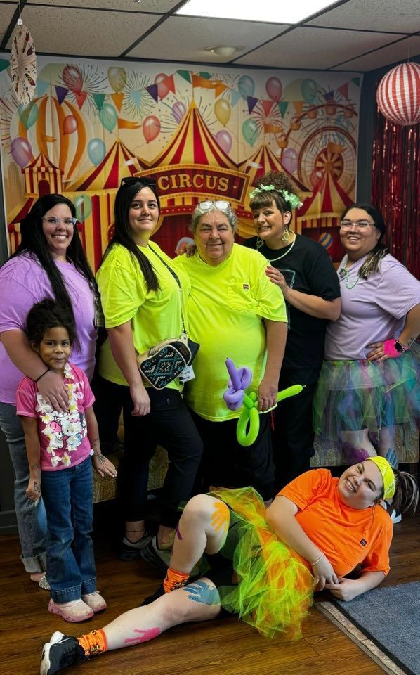 Group of people in circus-themed setting; some in bright clothes, one with face painted, posing near a