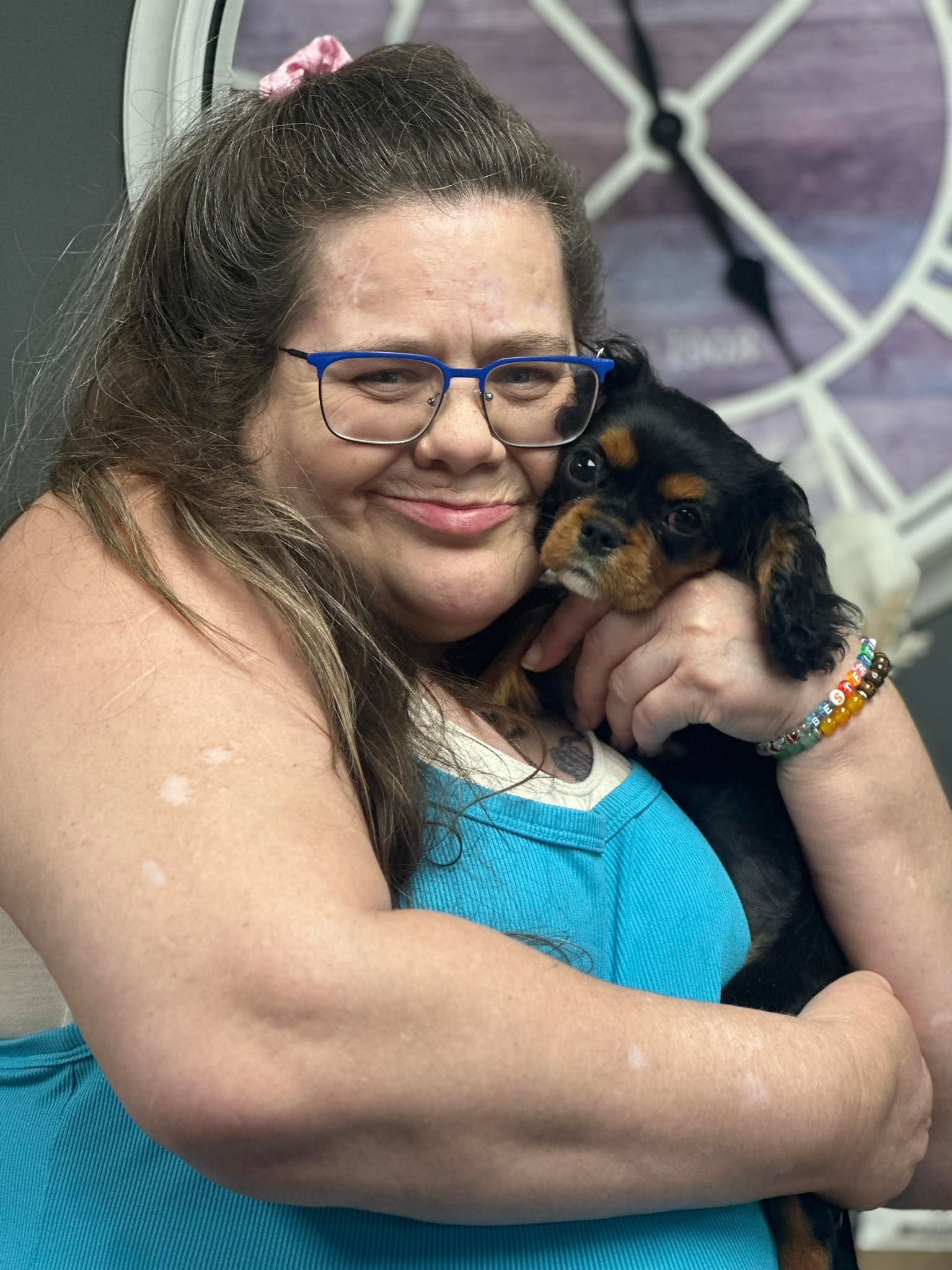 Woman in glasses holding a puppy, both smiling. Woman wearing blue tank top, pink scrunchie. Background: clock.