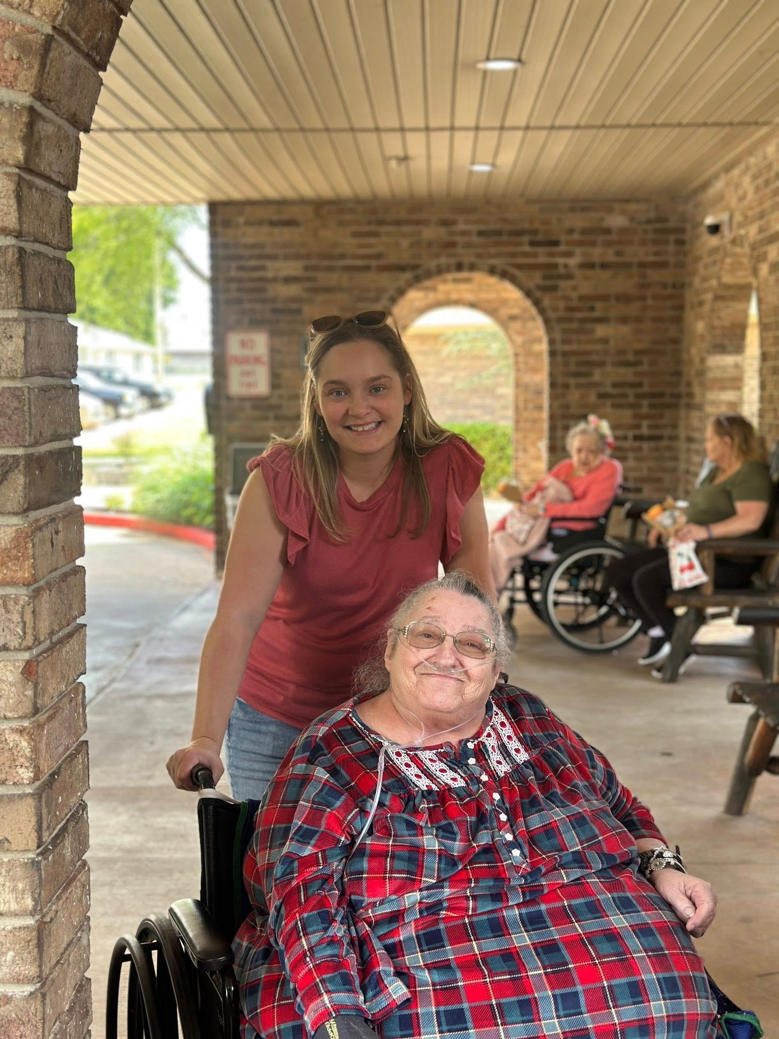 Woman pushing elderly woman in wheelchair; brick archway, outdoor setting.
