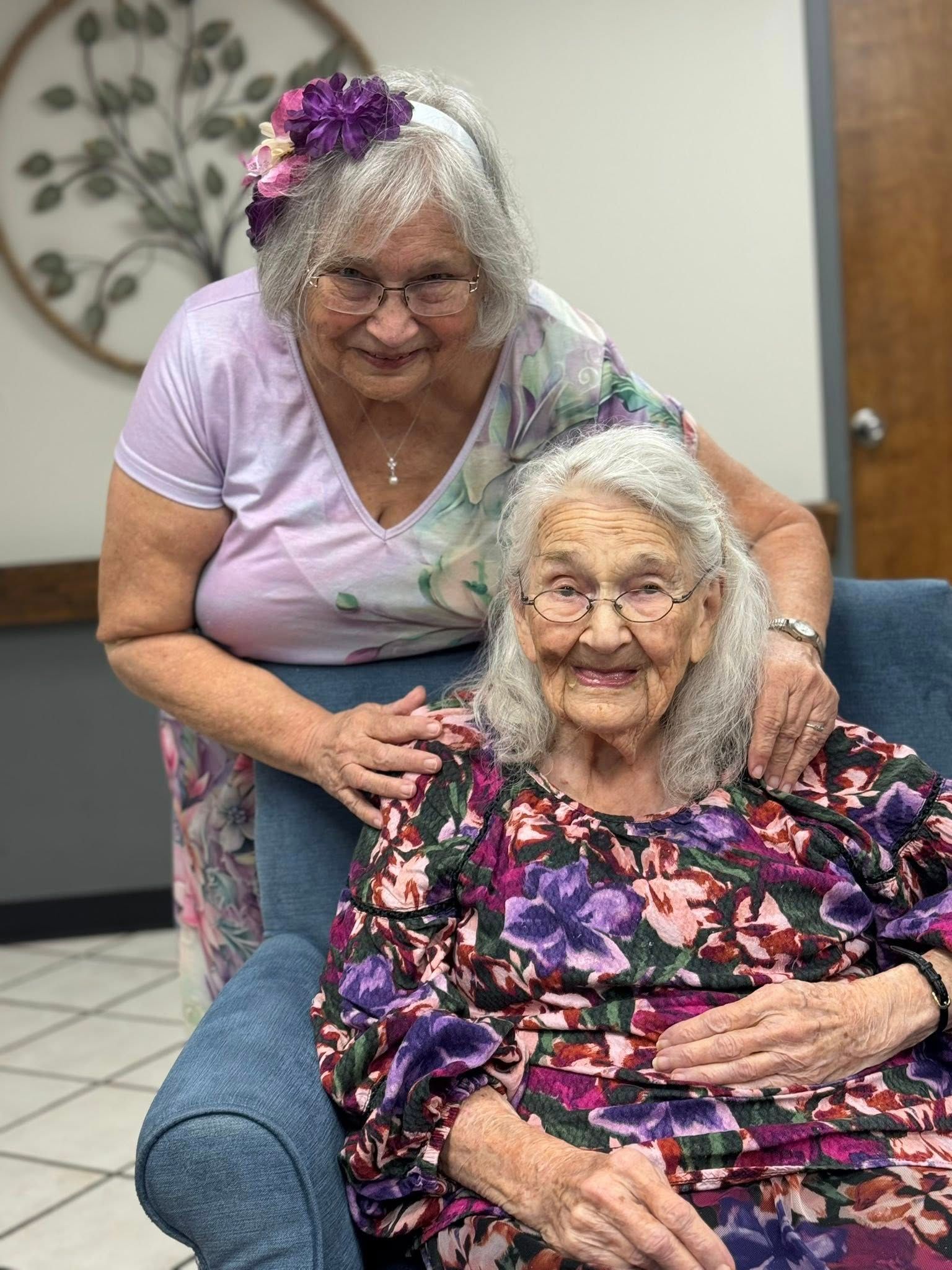 Two elderly women smiling: one seated, one standing behind her with a hand on her shoulder.