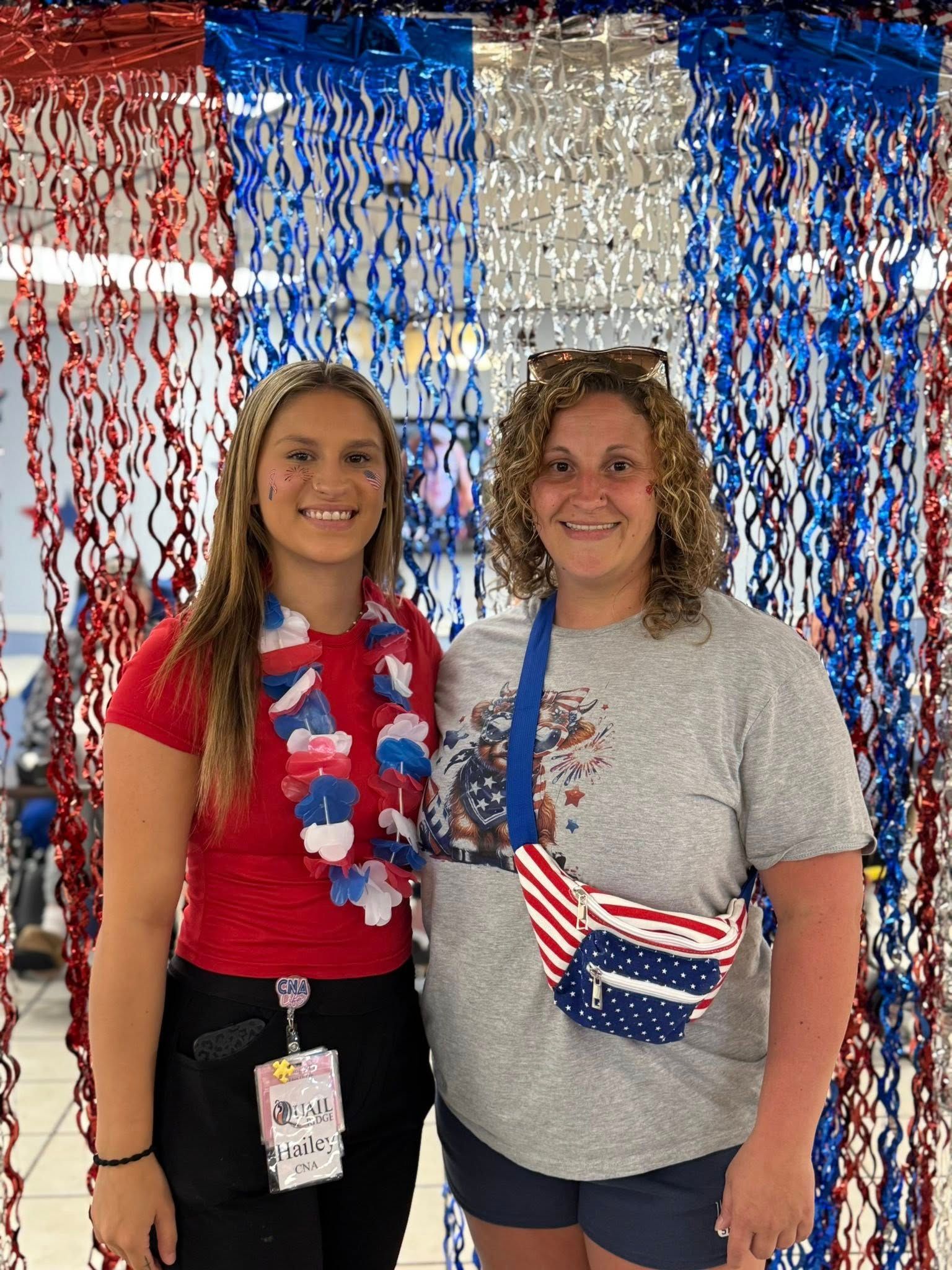 Two women pose in front of red, white, and blue streamers. One wears a red shirt, the other a gray shirt and fanny pack.