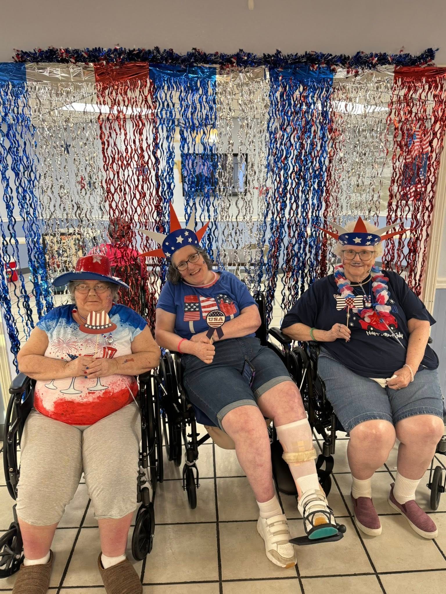 Three women in wheelchairs pose in front of patriotic decorations, wearing festive hats.