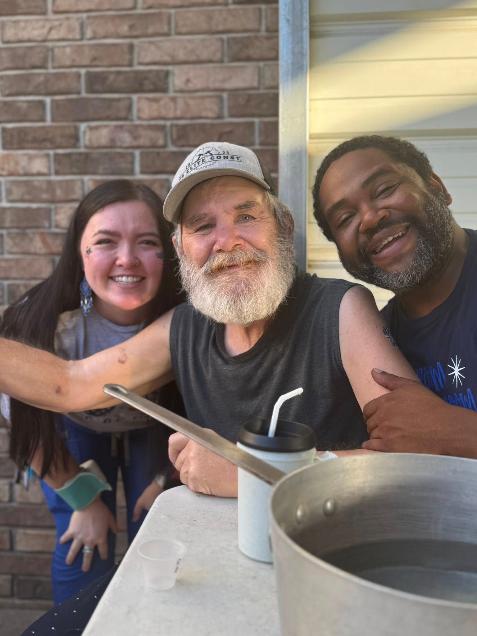 Three smiling people pose outdoors. A woman, a man with a beard, and a Black man. They stand near a table with a pot and drinks.
